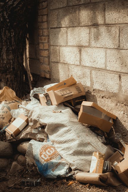 A large, weathered beige garbage bag made of thick, plastic material rests on the pavement beside a building wall in an urban alleyway. The bag appears full and bulging, with some items partially visi