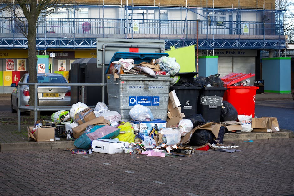 A large collection of mixed waste and rubbish bags is piled outside a recycling and waste collection point on a paved urban street. The trash includes black and black plastic bin bags, some opened, revealing paper, packaging, and miscellaneous rubbish. There are several cardboard boxes and paper-based materials scattered around, along with discarded packaging and flattened cartons. A gray metal recycling container designated for mixed paper and card is visible, with its lid open and overflowing with newspapers, cardboard, and paper waste. Nearby, a red plastic drum is present, possibly used for general waste. Behind the refuse pile, a car is parked on the street, and a low metal railing separates the waste area from the pedestrian walkway. In the background, there are storefronts with signage, a storefront with a sign indicating a fish bar, and a building with blue scaffolding and safety barriers, suggesting ongoing work. The scene appears to be in a busy urban environment where rubbish removal services like those by Rubbish Removal London are employed for waste management, especially in cases of large, unsorted rubbish accumulations typical of private collection or on-site clearance.