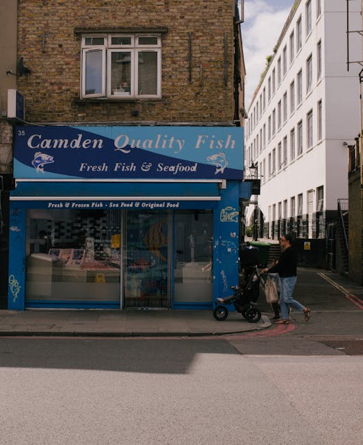 The image depicts the facade of a small fish and seafood shop called 'Camden Quality Fish,' with a blue sign displaying the shop's name in white lettering and images of fish. The shop has a large glass display window showing various fresh and frozen seafood products inside, with a white countertop visible beneath the window. Above the shop, there is a brick building with a white-framed window, partially obscured by the shop sign. Next to the shop, on the right, there is a modern multi-storey building with white exterior walls and multiple windows. On the pavement in front of the shop, a person dressed in dark clothing is pushing a black stroller with a shopping bag hanging from it, appearing to be engaged in a casual activity. The scene is set during the day with natural daylight illuminating the area, and the overall environment appears urban, with the surroundings indicating a busy street or commercial area. This setting may be linked to private waste disposal or rubbish removal services, such as those provided by Rubbish Removal London, especially given the proximity to commercial establishments that might require such services.