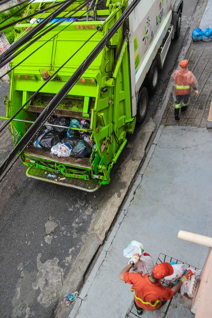 A bright green refuse collection truck is parked on a narrow urban street, with its rear compartment open, revealing a load of black plastic rubbish bags filled with waste. The truck's surface has a s