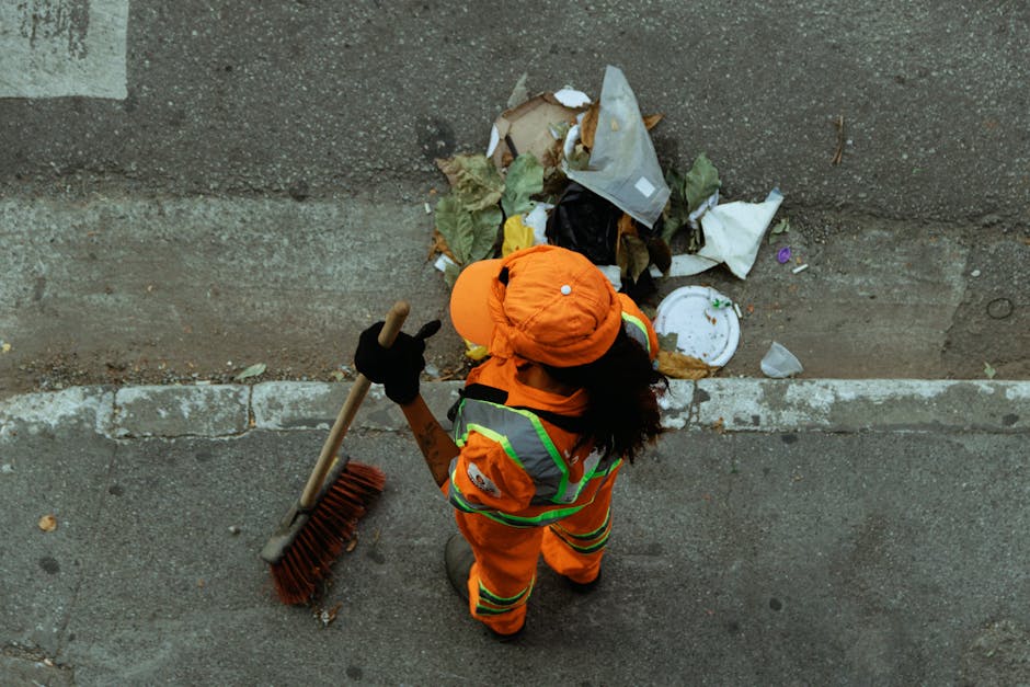 A bright green refuse collection truck is parked on a narrow urban street, with its rear compartment open, revealing a load of black plastic rubbish bags filled with waste. The truck's surface has a s